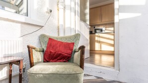 Un fauteuil vert avec un coussin rouge, placé dans un coin d’où l’on aperçoit une cuisine et une fenêtre.