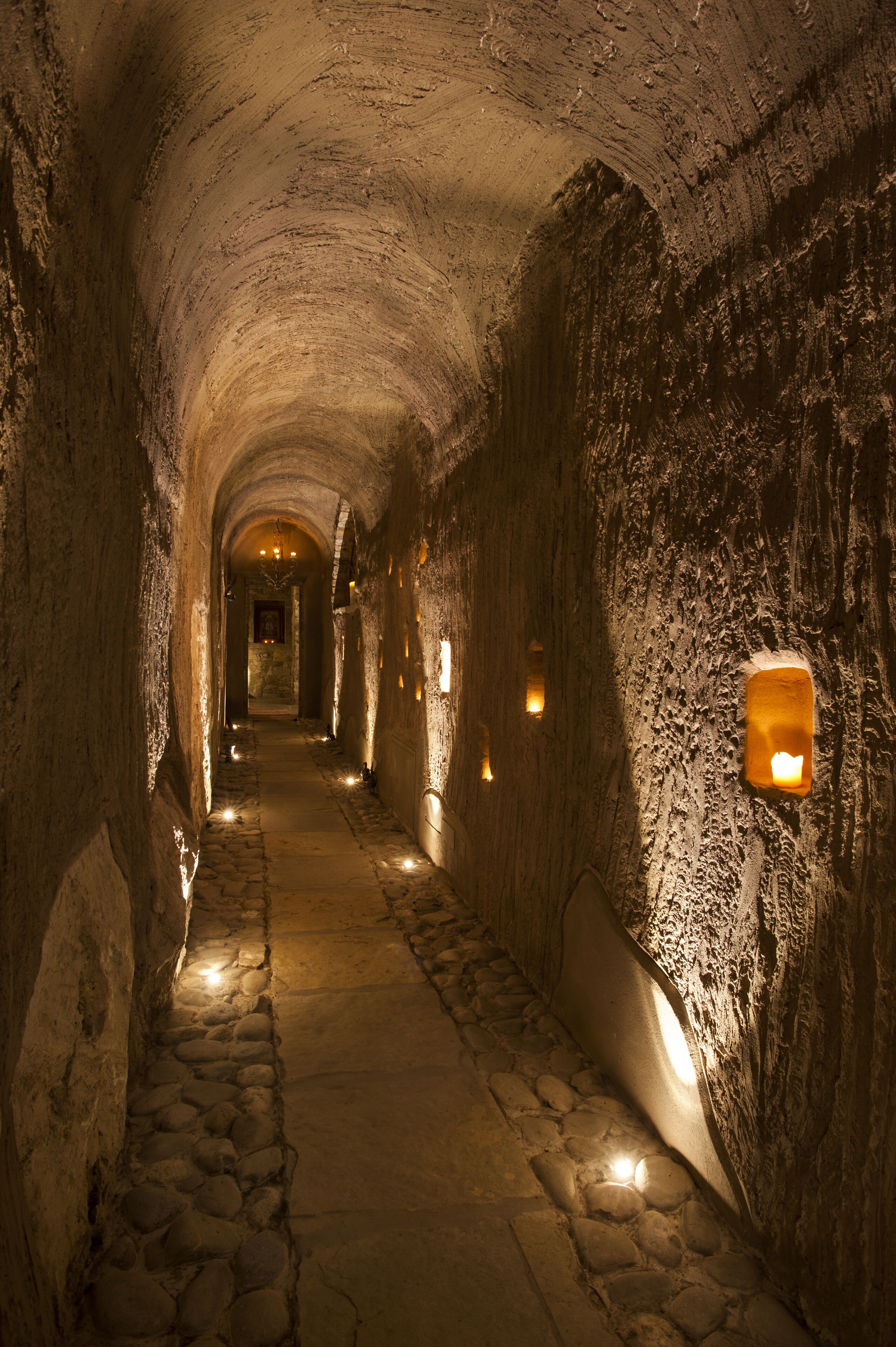 A stone tunnel with a narrow path, illuminated by wall-mounted lights.