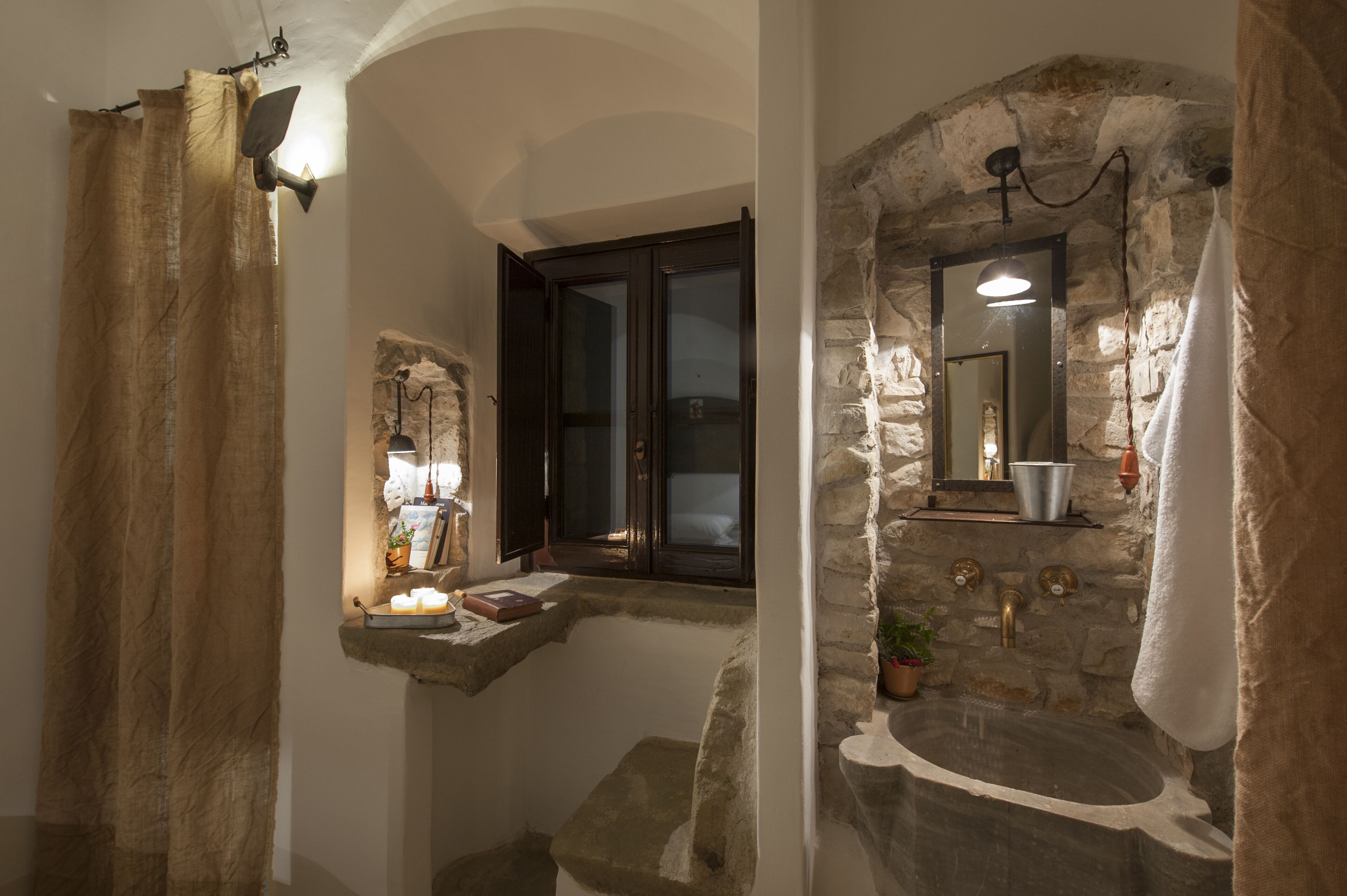 A bathroom with stone walls, a window with dark shutters, a sink with a mirror, and a towel hanging on the wall.