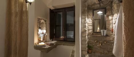 A bathroom with stone walls, a window with dark shutters, a sink with a mirror, and a towel hanging on the wall.