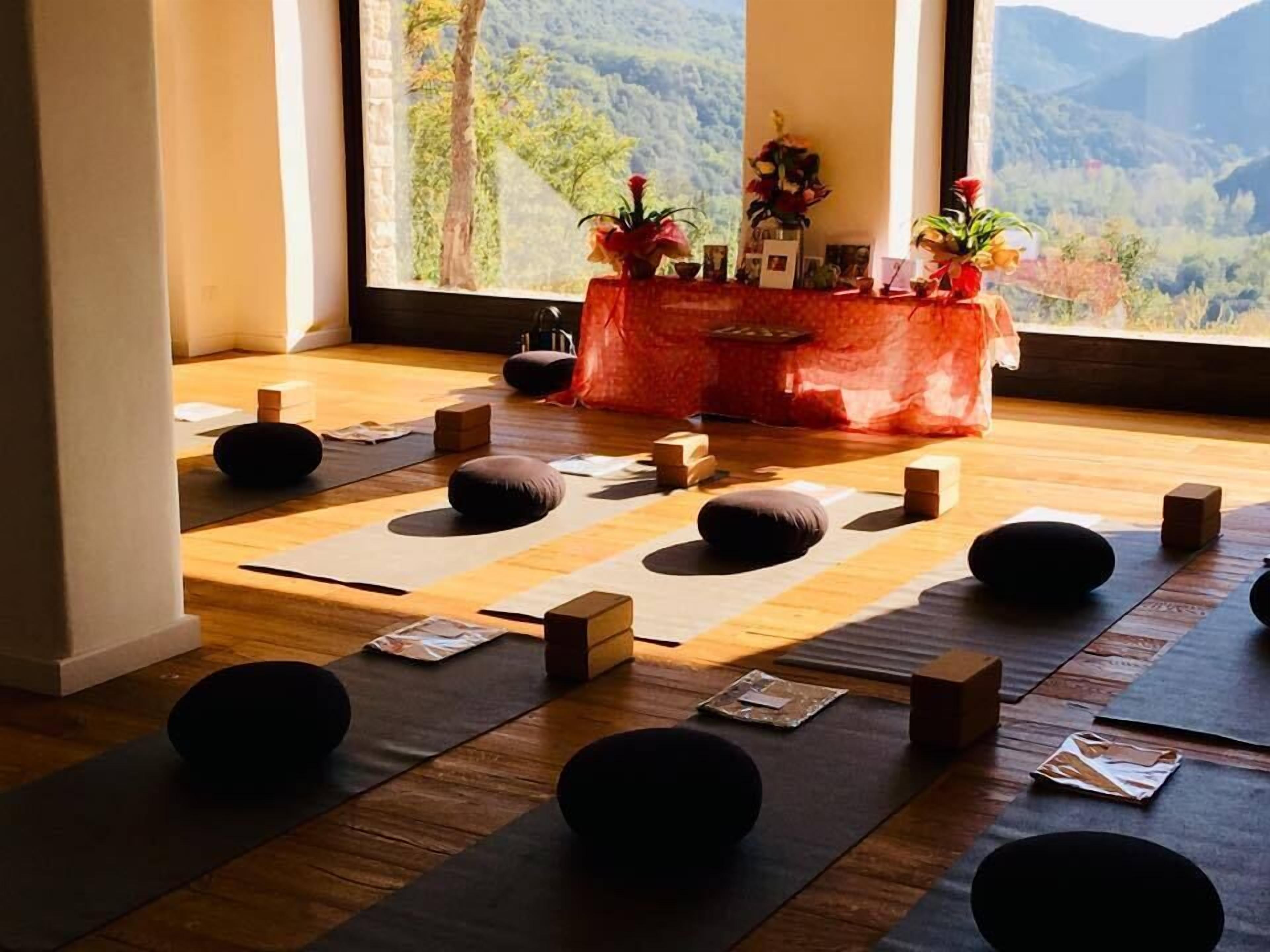 Une pièce dotée d’un plancher en bois, de tapis noirs et de blocs de bois disposés pour une séance de yoga ou de méditation. Une fenêtre offre une vue sur un paysage montagneux.
