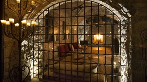 A stone-walled room with a bed, a chandelier, and a view through a barred window. - Eremito (Parrano)