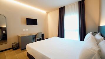 Habitación de hotel con cama, escritorio, silla, televisor y ventana con cortinas.