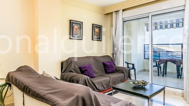Ein modernes Wohnzimmer mit einer Couch und Sesseln, einer Glastür, die auf einen Balkon führt, und gerahmten Kunstwerken an der Wand.