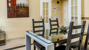 A dining area with a glass table and four chairs, a painting on the wall, and an open door leading to another room.