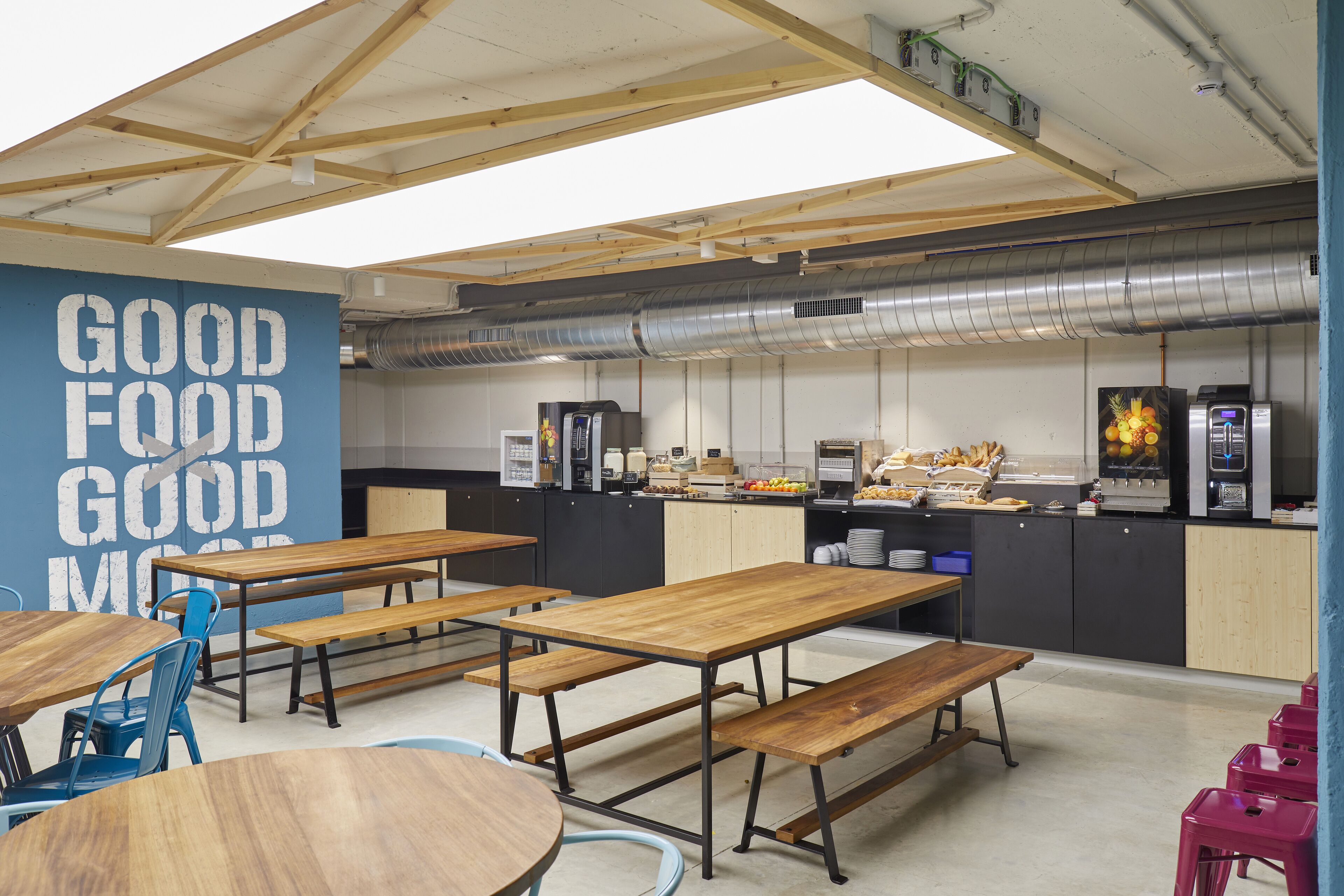 A modern dining area with wooden tables and benches, a kitchen in the background, and a blue wall with the text "GOOD FOOD GOOD MORNING".
