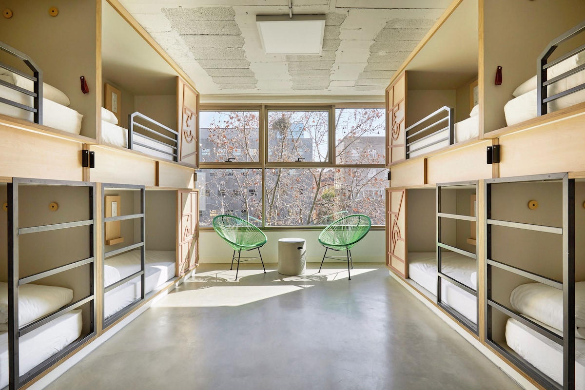 A dormitory room with bunk beds, a large window, and two green chairs.