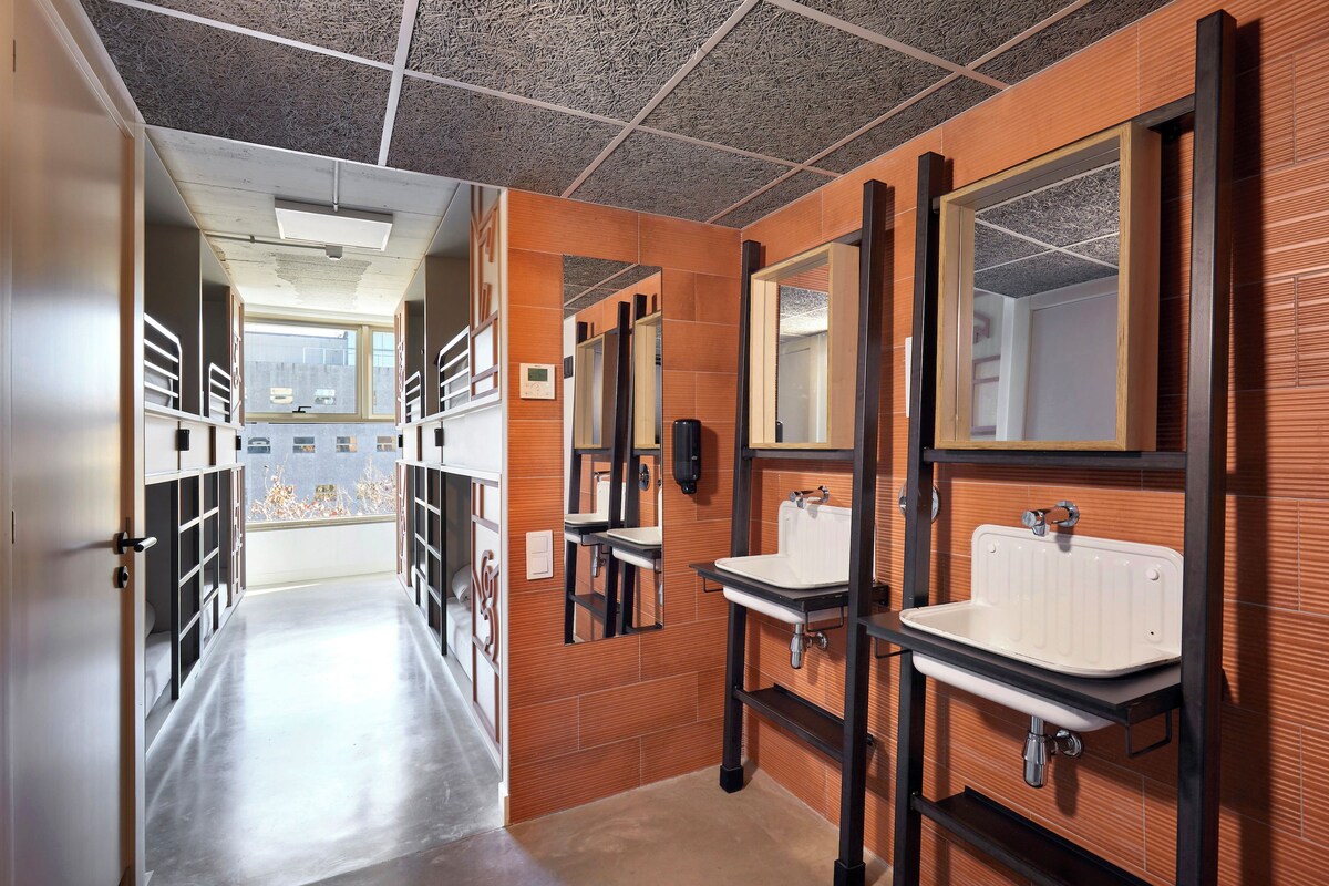 A corridor with multiple mirrors and sinks, leading to a window with a view of buildings.