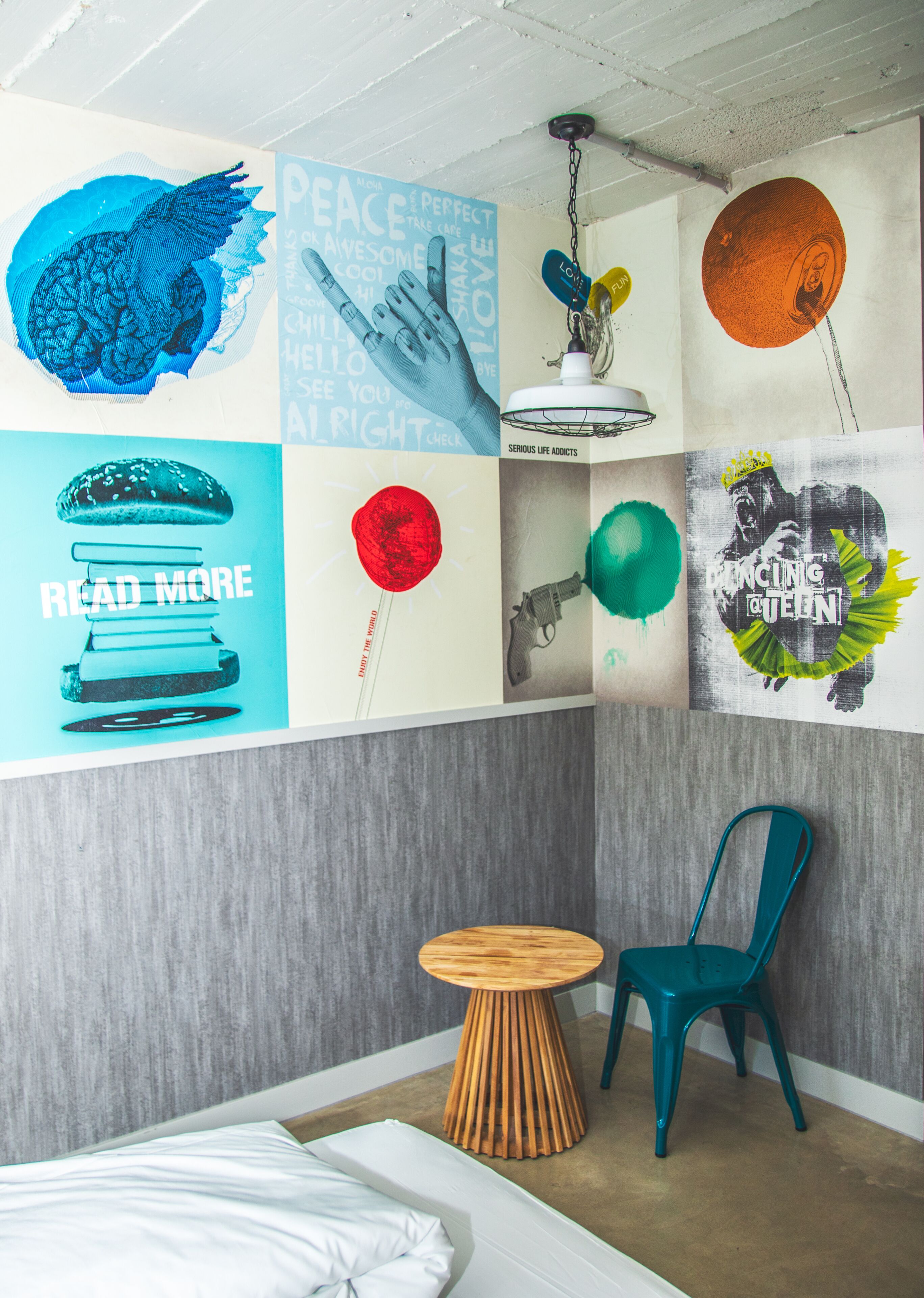 A room with a bed, a teal chair, a small wooden table, and various wall art including a blue bird, a red lollipop, and a poster with a crown.