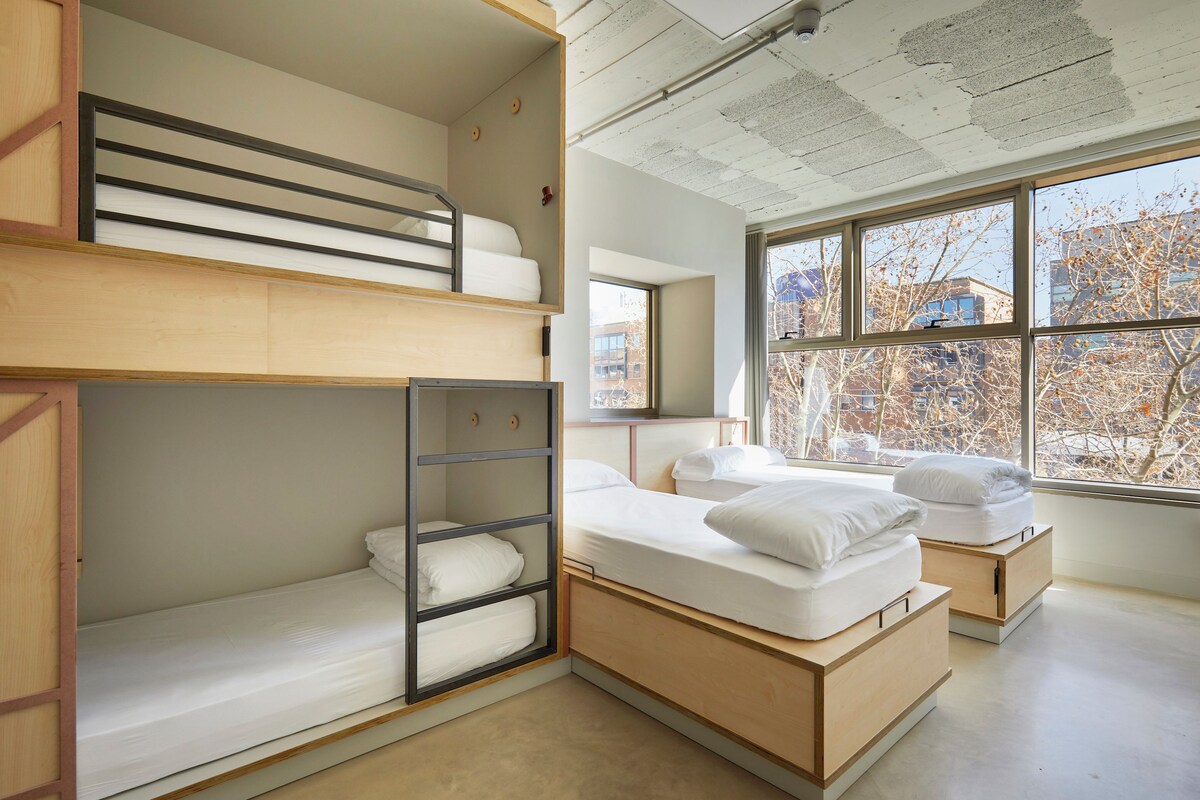 A modern, multi-level dormitory room with bunk beds, built-in storage, and large windows offering a view of the city.