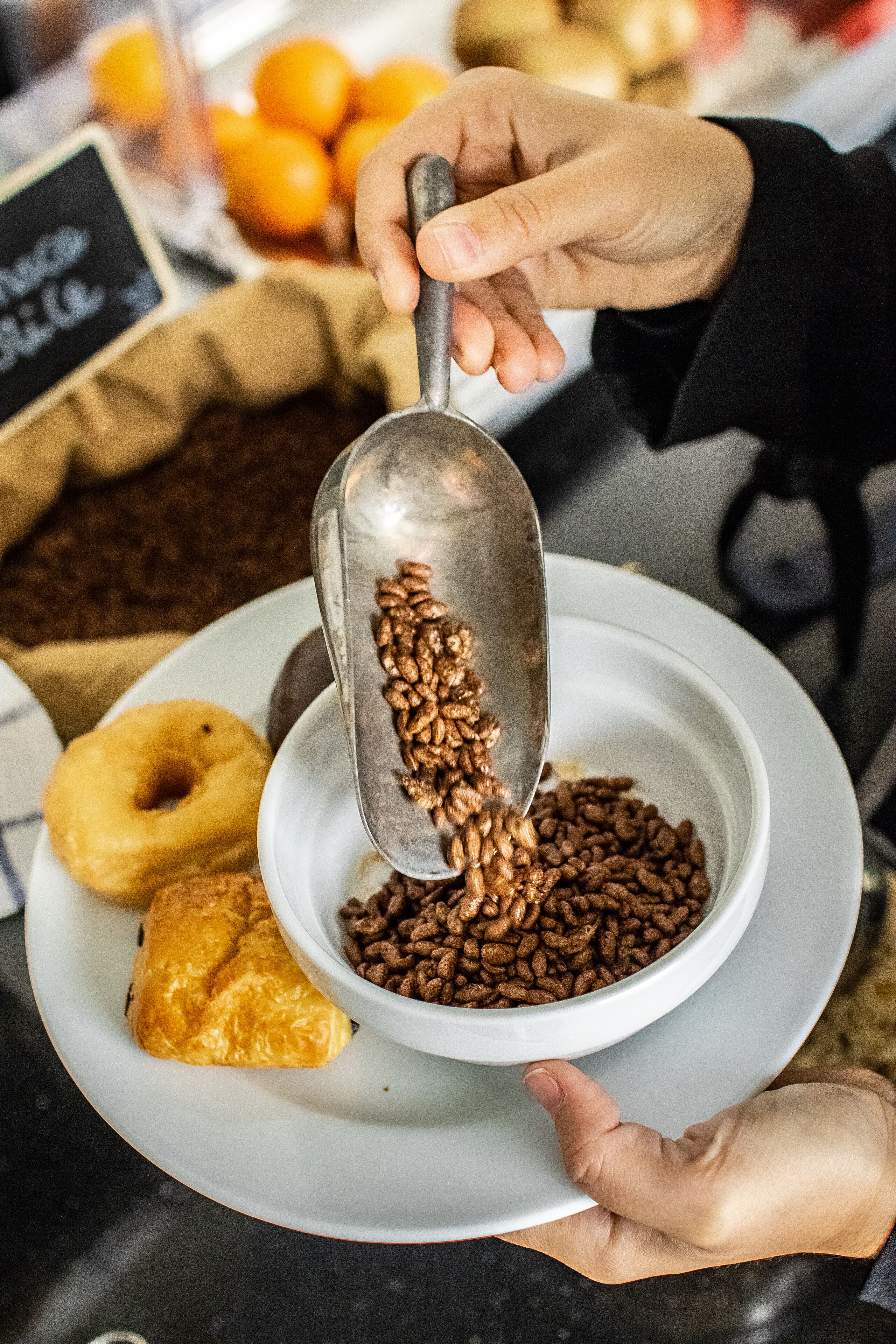 Une personne verse des grains de café dans un bol, avec des pâtisseries et des oranges en arrière-plan.