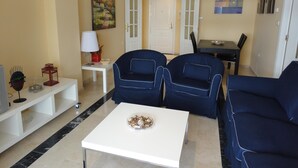 Un salon avec une table blanche, deux fauteuils bleus, une petite table d’appoint blanche et un tableau accroché au mur.
