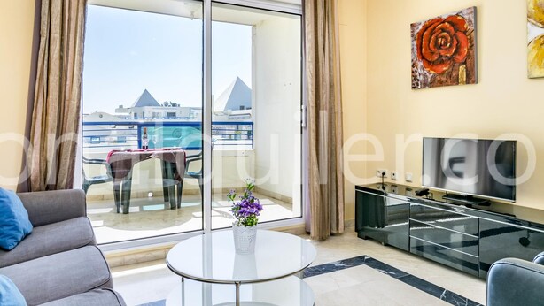 Sala de estar moderna com mesa de vidro, sofá, televisão e vista para uma piscina e edifícios.