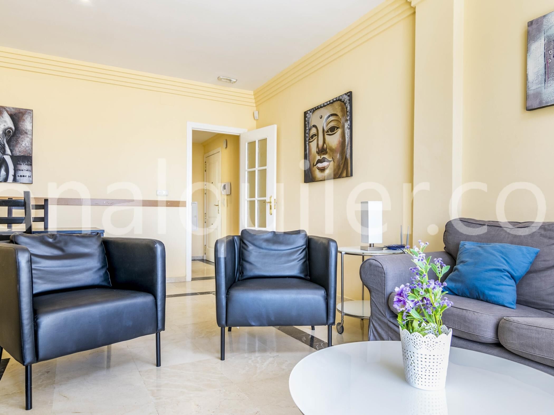 Un salon moderne avec deux fauteuils en cuir noir, un canapé gris et une table basse blanche ornée d’un vase rempli de fleurs violettes.