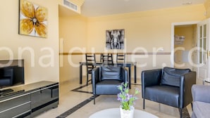 Uma sala de estar moderna com televisão de tela plana, um espaço de jantar com mesa e cadeiras, e uma mesa de centro branca com um vaso de flores.