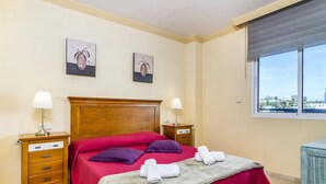 A hotel room with a wooden headboard, two bedside tables with lamps, a bed with red and purple bedding, two wall-mounted artworks, and a window with a city view.