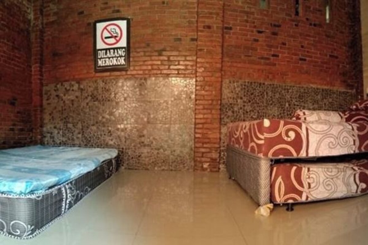A room with two beds, one with a blue mattress and the other with a patterned cover. A no-smoking sign is visible on the wall.