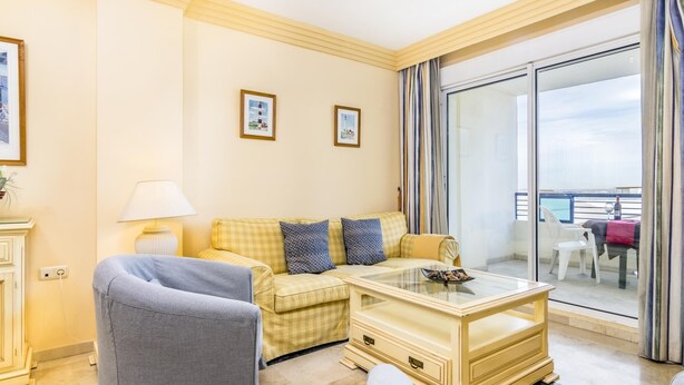 A hotel room with a checkered sofa, a glass coffee table, a grey armchair, and a view of the balcony through a large window. - Magnifico apartamento en Marbella con vistas al mar (Marbella)