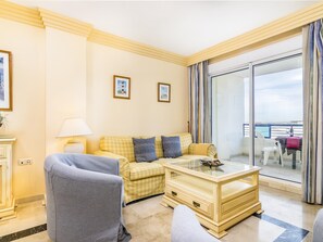 Habitació d'hotel amb un sofà estampat de quadradets, una taula de cafè de vidre, un armari gris i vista al balcó a través d'una finestra gran.
