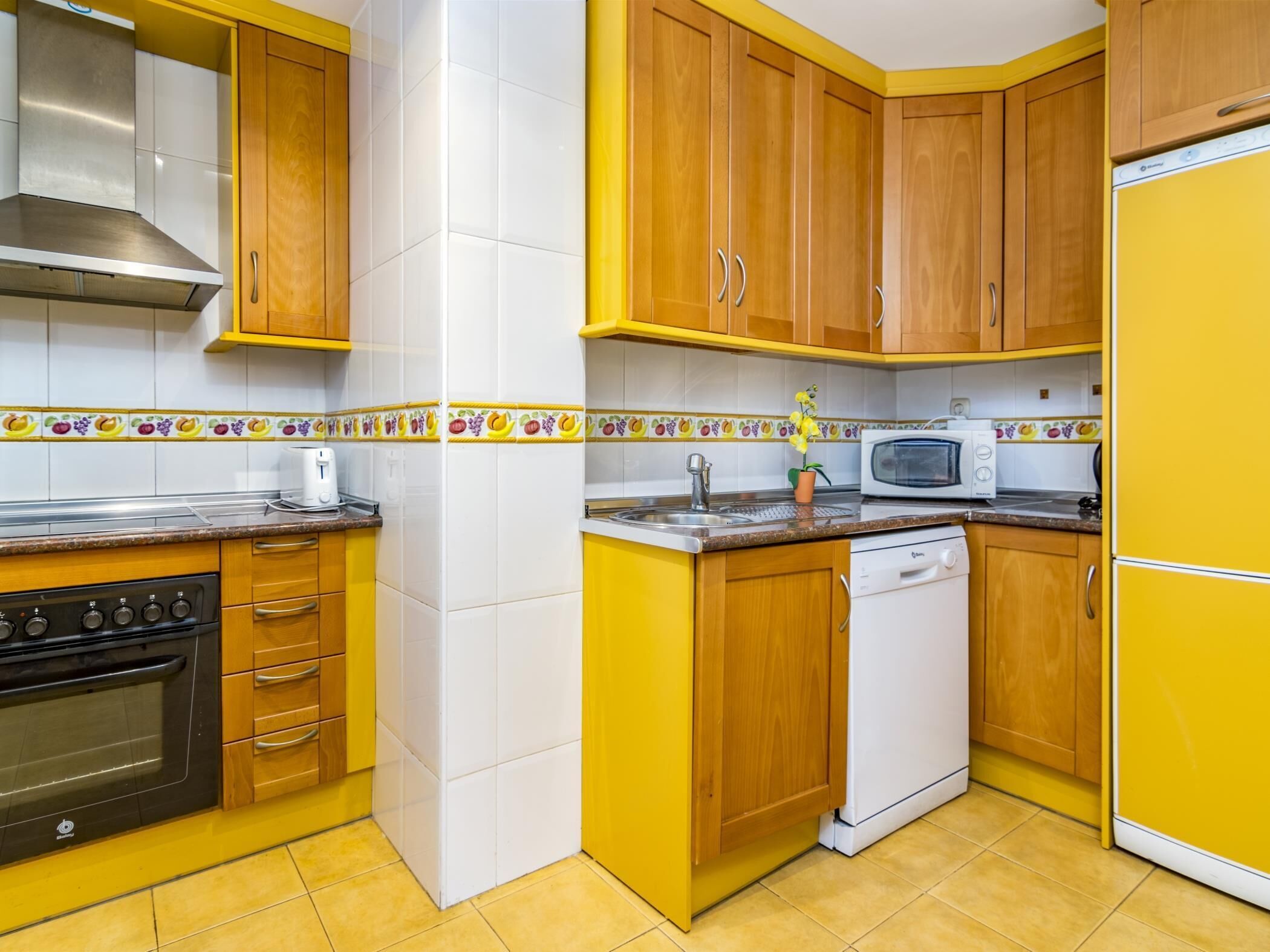 A kitchen with yellow cabinets, a stainless steel range hood, a microwave, a dishwasher, and a toaster.