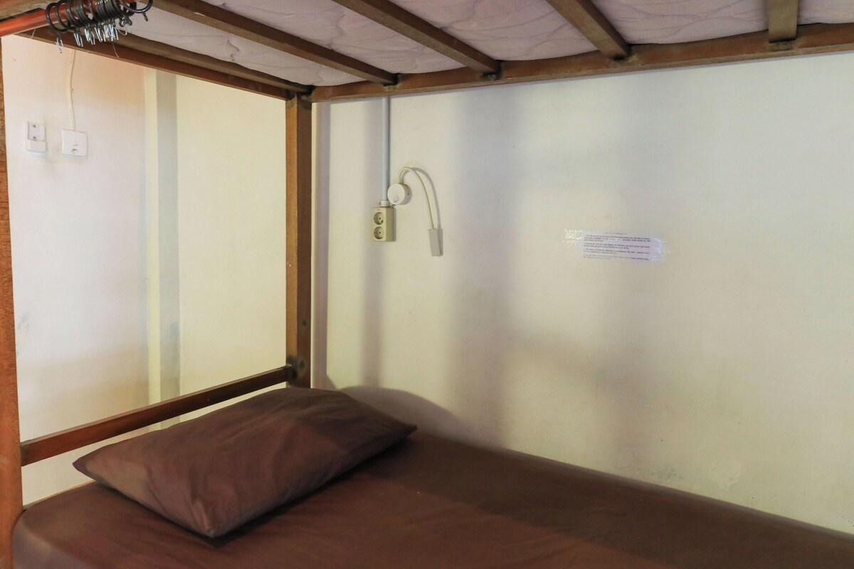 A single bed with a brown mattress and pillow, a window, and a wall-mounted electrical outlet.