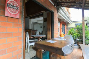 A brick exterior with a wooden table and chairs, a sign for Bintang Pilsener beer, and a view of the street. - Halaman Depan Hostel (Ubud)