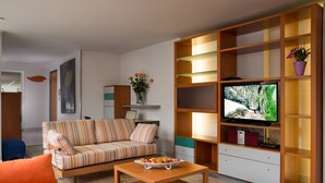 Un salon avec un canapé à rayures, une table basse en bois, un téléviseur posé sur un meuble en bois et une bibliothèque agrémentée d’une plante en pot.