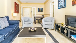 A living room with a blue sofa, two grey armchairs, a coffee table, a TV on a black stand, and a dining area in the background. - Magnifico apartamento en Marbella con vistas al mar (Marbella)