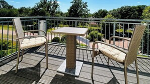 A balcony with wicker furniture, a table, and a view of the surrounding greenery and buildings. - Hotel Reindahls (Altlandsberg)