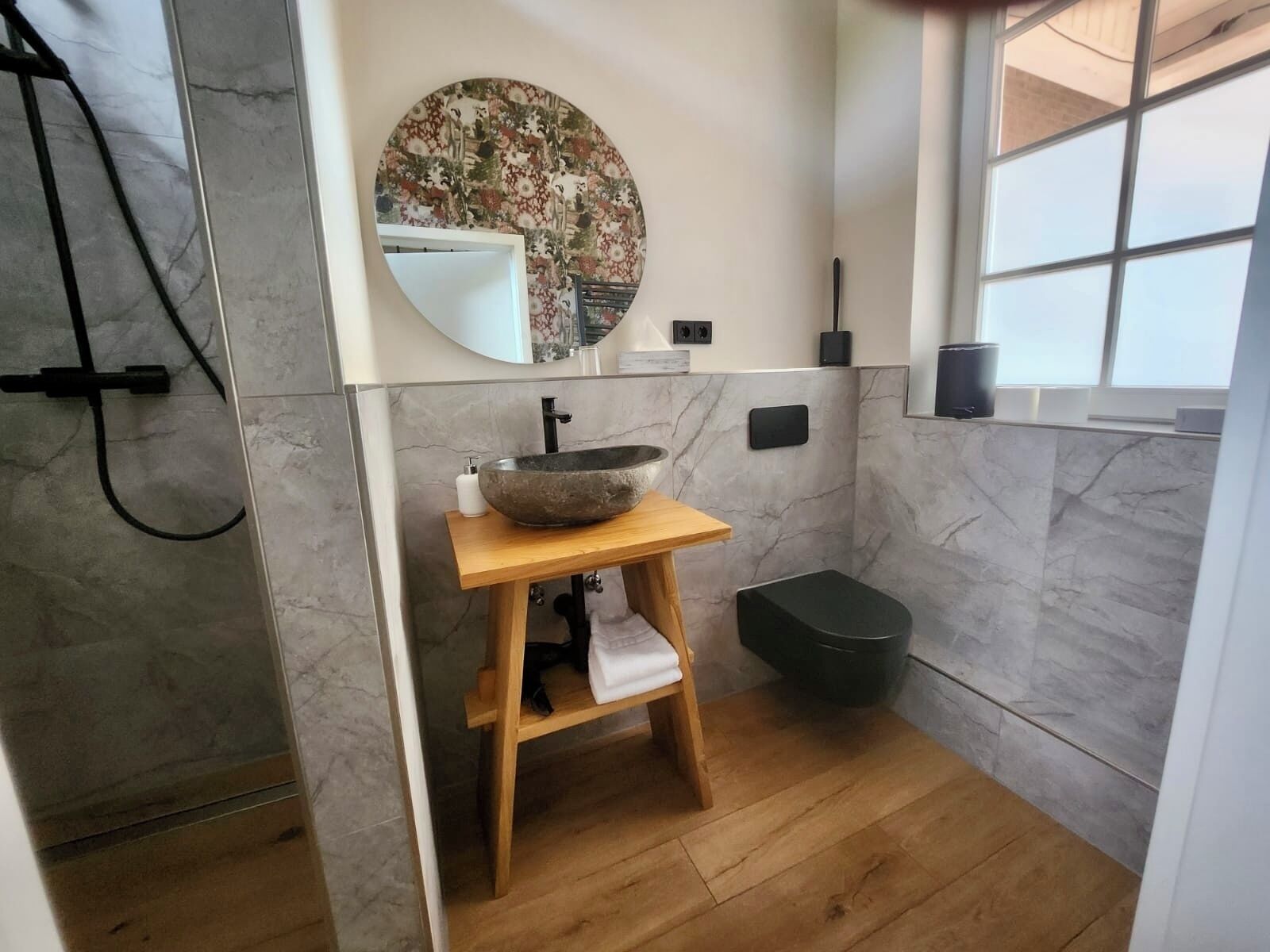 A bathroom with a wooden vanity, a large round mirror, a stone basin, a black trash bin, and a shower area.