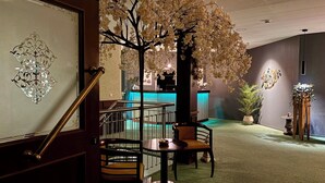 A lobby with a reception desk, a staircase with a handrail, a round table with chairs, and a decorative tree with white flowers. - Hotel Reindahls (Altlandsberg)