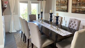 A dining room with a dark wooden table, beige upholstered chairs, and a patterned table runner. There are two tall, dark blue candle holders with white candles on the table. The room has a large window with white curtains, and a painting is visible on the wall. - Elegant West Side Mountain Retreat (Tucson)