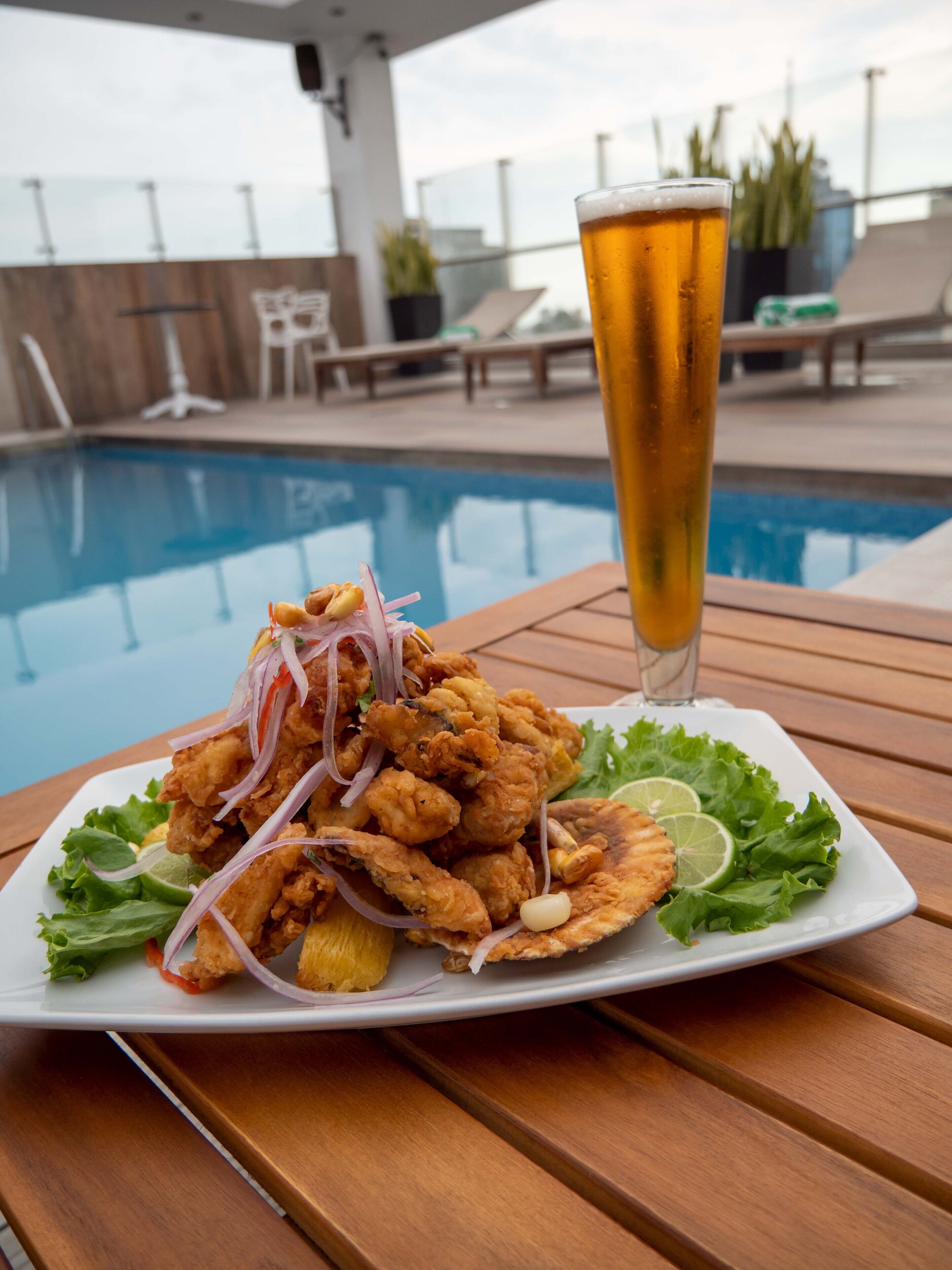 Plat de menjar fregit amb un got de cervesa sobre una taula de fusta al costat d'una piscina.