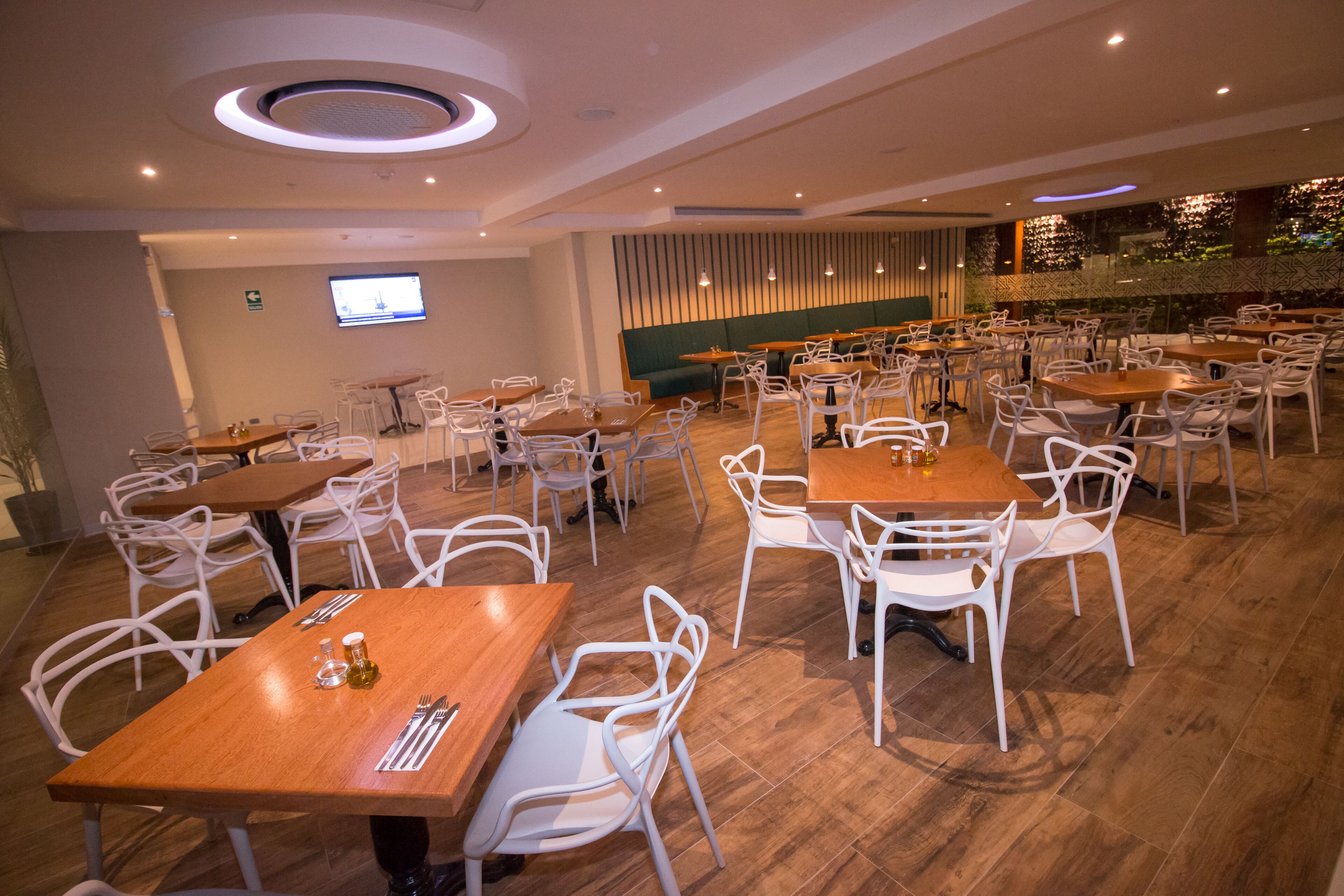 Um restaurante espaçoso com várias mesas e cadeiras, uma tela grande na parede e um espaço com bancos acolchoados verdes.
