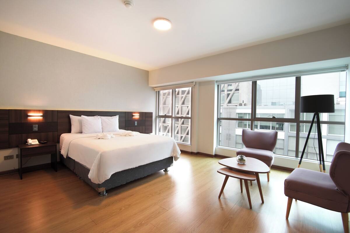 Ein Hotelzimmer mit Bett, Nachttisch, Lampe, Sessel und Blick auf die Stadt durch große Fenster.