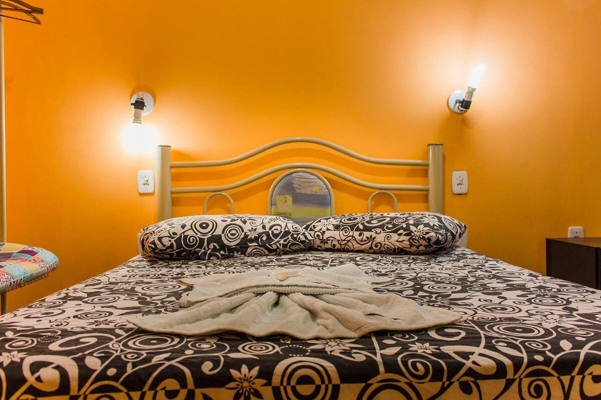 A bed with a patterned bedspread and pillows, a headboard with a light fixture, and a wall socket.