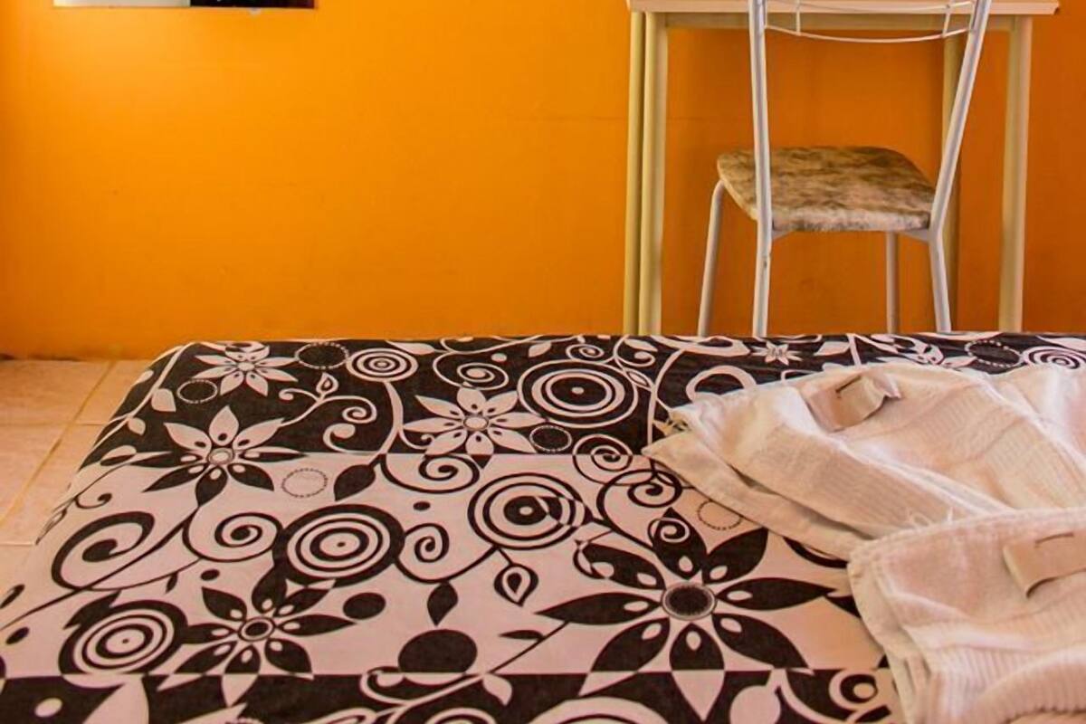 A room with an orange wall, a bed with a patterned bedspread, a small table with a chair, a mirror, and a mounted television.