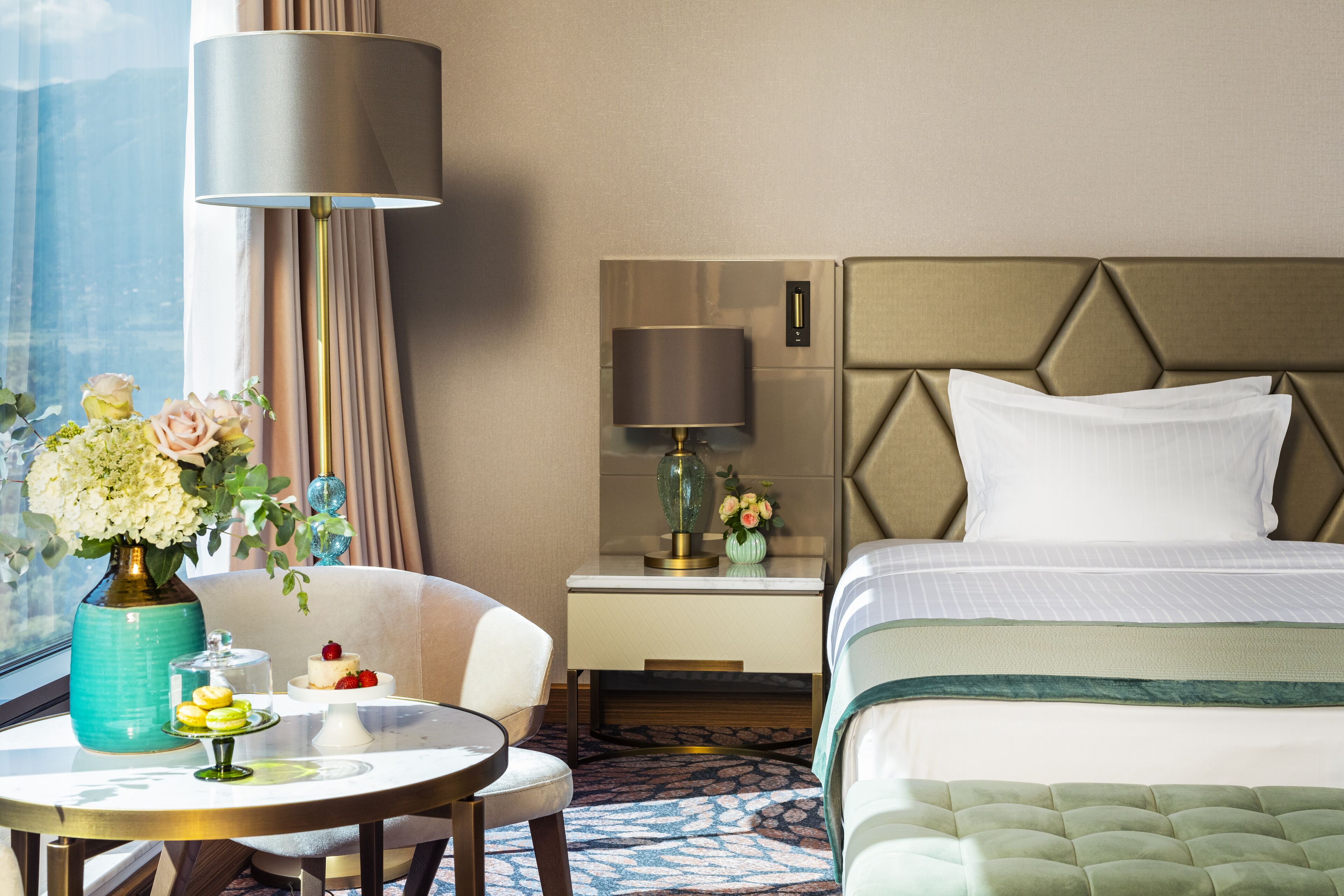 A hotel room with a bed, a bedside table with a lamp, a chair, a small table with a vase of flowers, and a view of the outdoors.