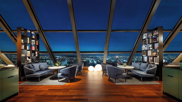 A modern interior with a panoramic city view, featuring a wooden floor, two sofas, a round table, and two bookshelves.