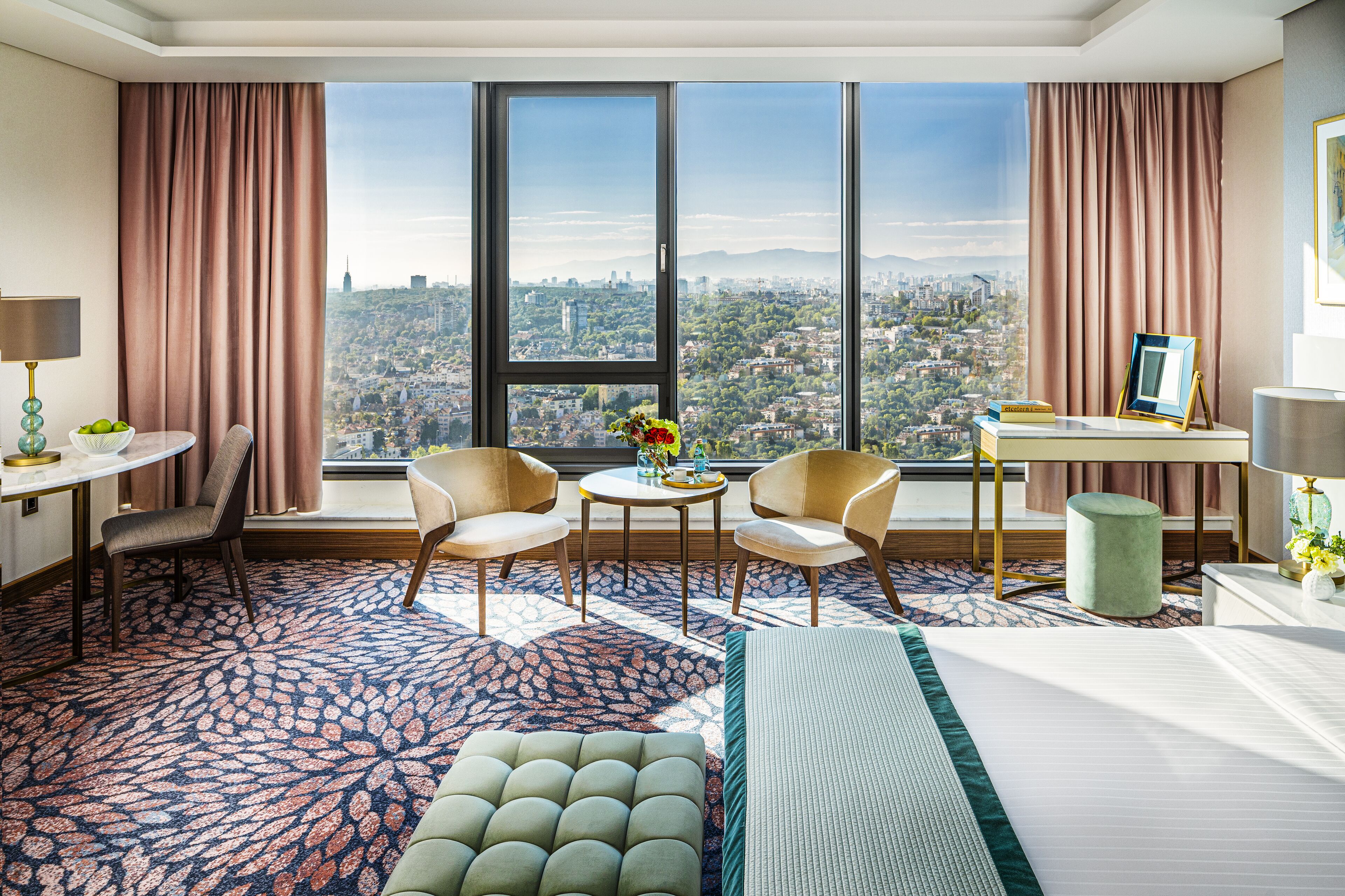 Habitación de hotel con un ventanal grande, una cama, un escritorio, dos sillas y una mesita con un jarrón de flores.