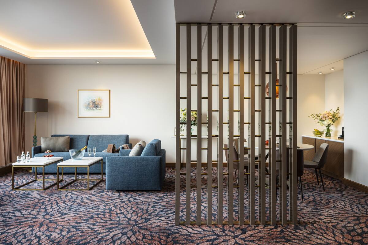 Une chambre d’hôtel moderne avec un canapé bleu, deux tables basses blanches, une moquette à motifs, un élément de séparation décoratif et un coin repas comprenant une table et des chaises.