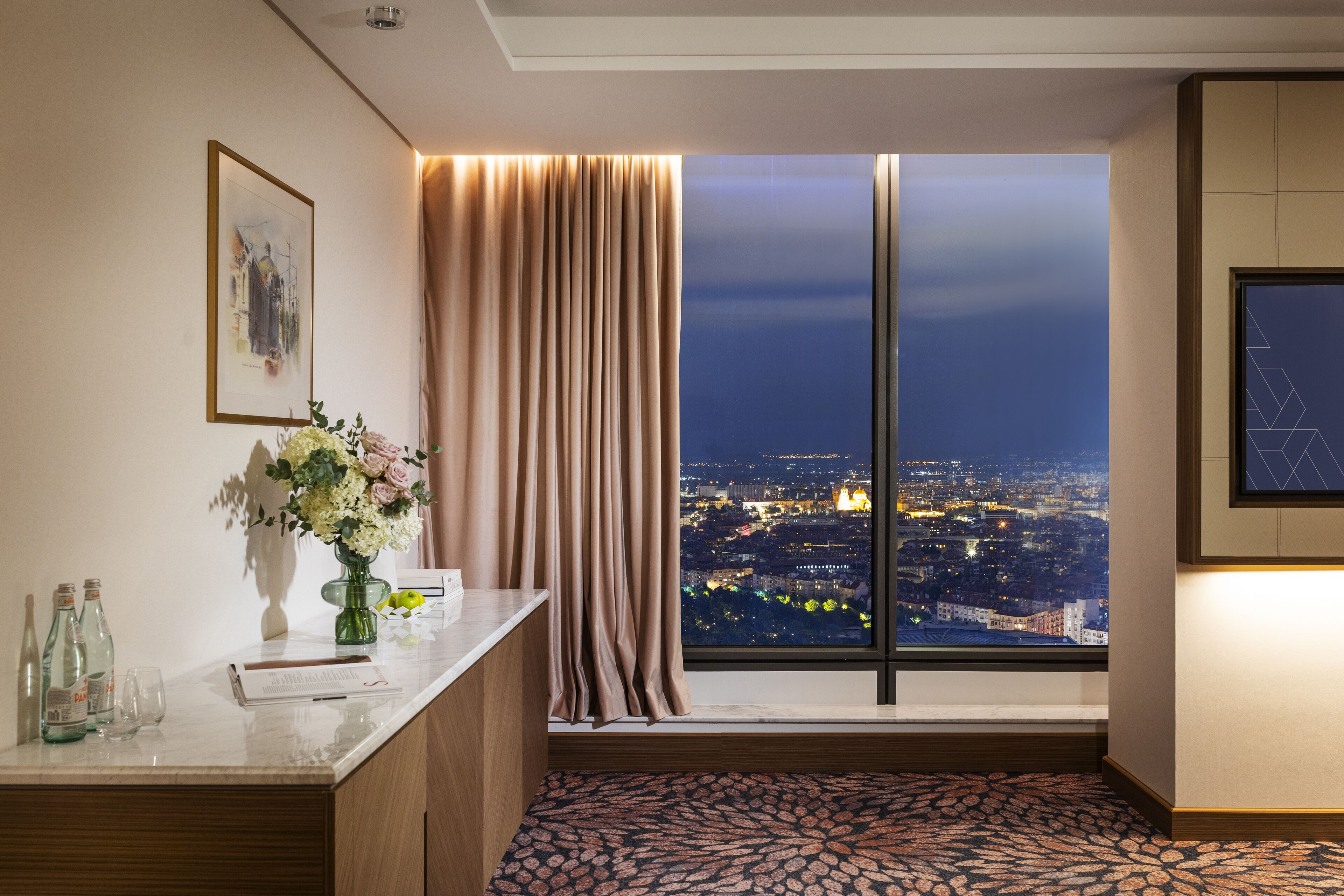 A hotel room with a city view, a desk with a vase of flowers, and a patterned carpet.