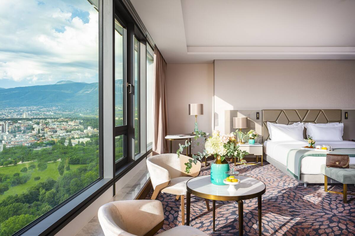 Une chambre d’hôtel avec une grande fenêtre, un lit, une table ronde ornée d’un vase de fleurs et une chaise.