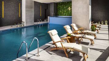 A modern indoor pool area with clear water, wooden lounge chairs, and a green wall with foliage.