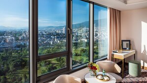 A hotel room with large windows offering a city view, a small round table with a vase of flowers, two chairs, and a desk with a chair.