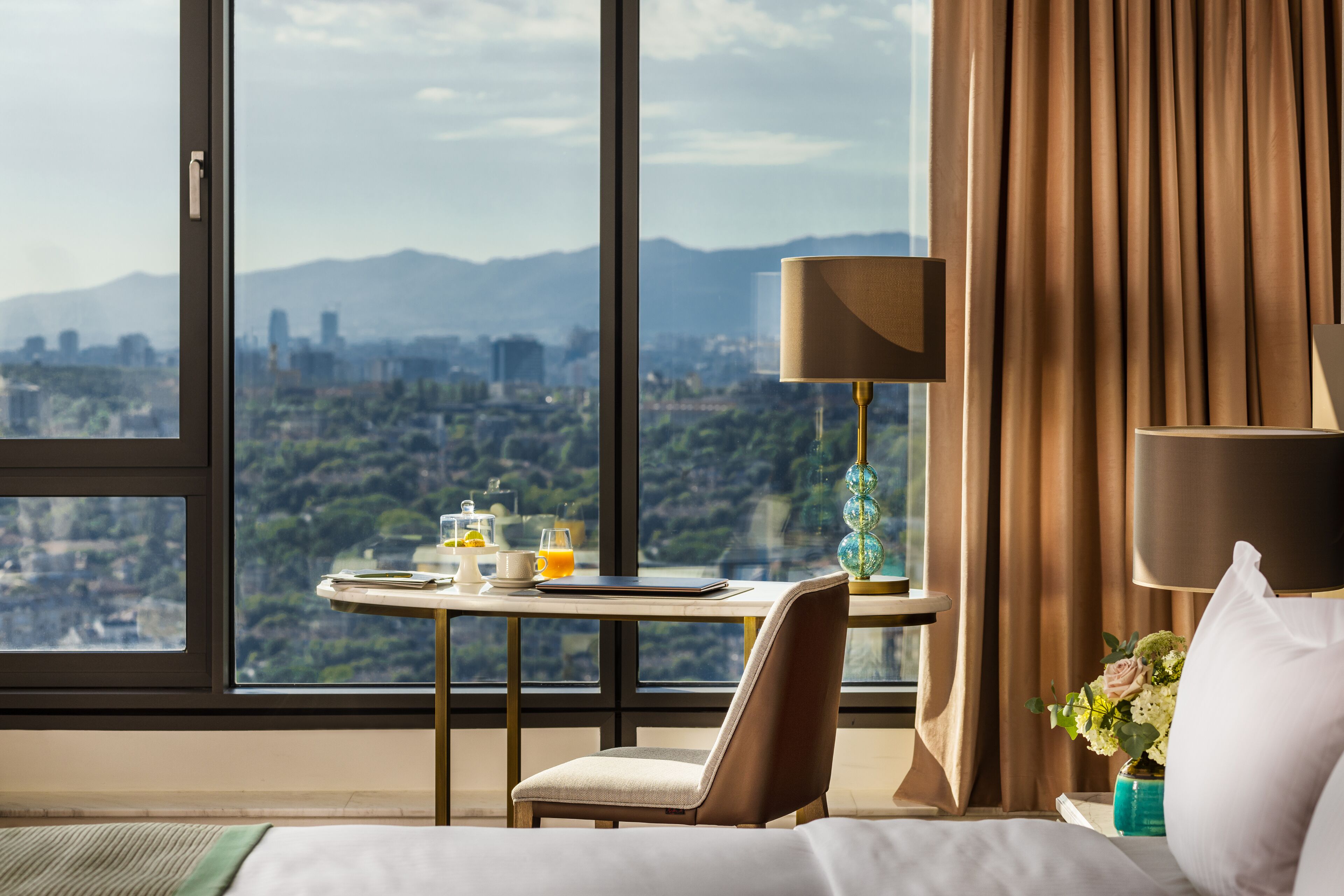 Ein Hotelzimmer mit großem Fenster und Blick auf die Stadt, einem Nachttisch mit Lampe und einem Sessel.