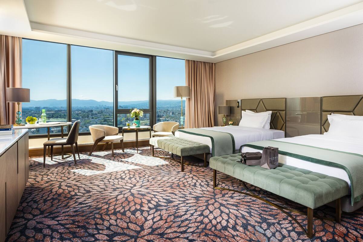 Une chambre d’hôtel avec une grande fenêtre, un bureau, deux lits et une moquette à motifs.