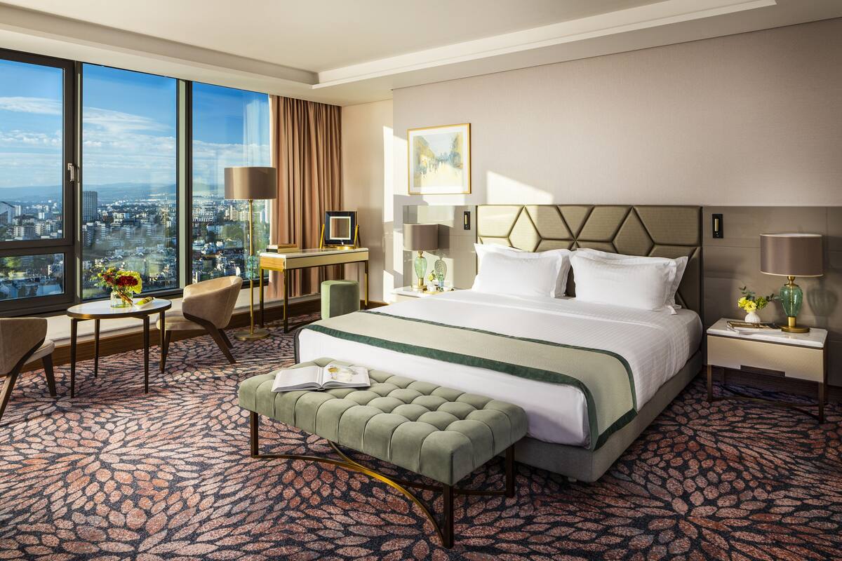 Une chambre d’hôtel avec une grande fenêtre, un lit recouvert d’une couverture verte et blanche, un bureau avec une lampe, une chaise et une petite table agrémentée de fleurs.