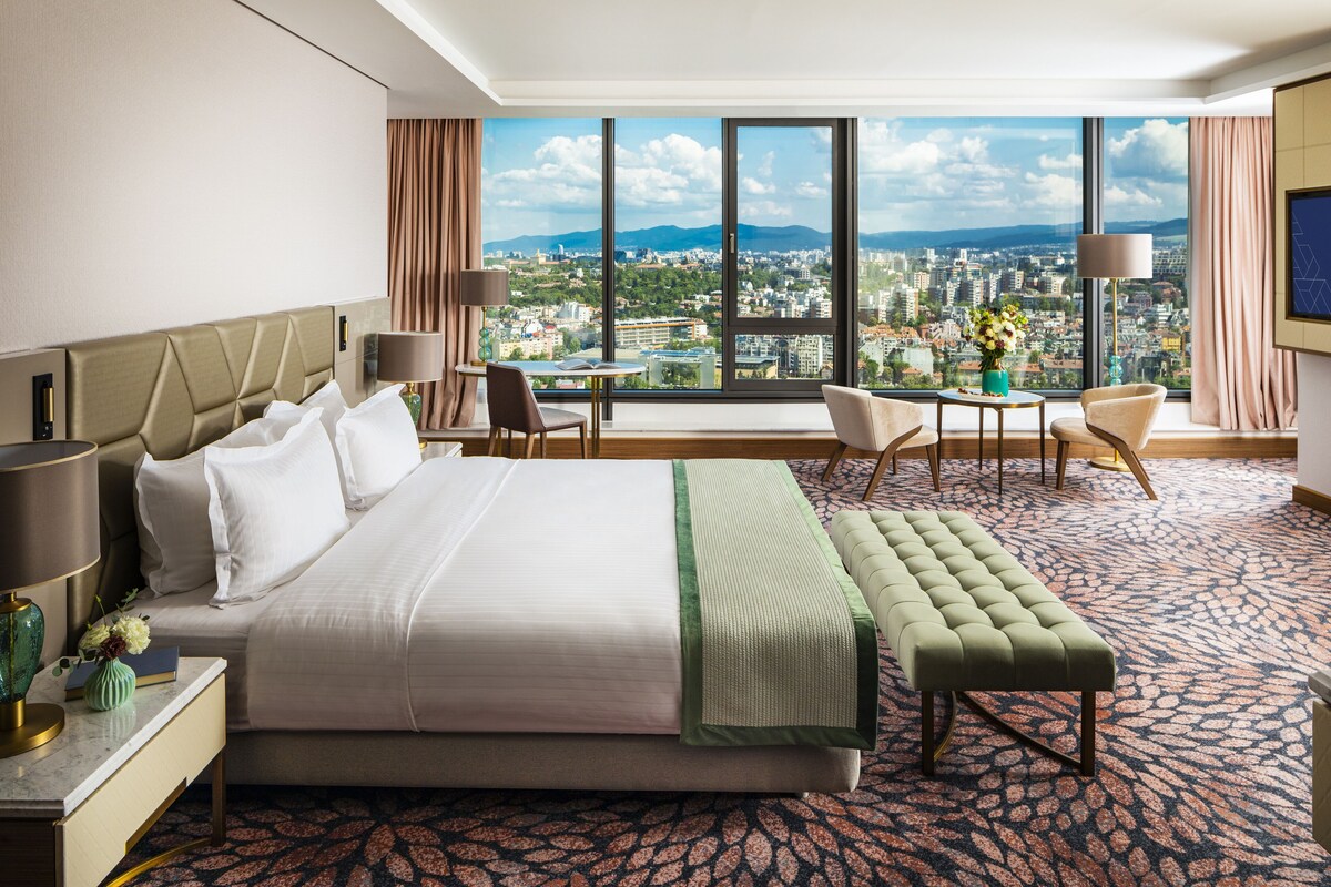 Une chambre d’hôtel avec un grand lit, un bureau, une chaise, une petite table et une vue sur le paysage urbain grâce à de grandes fenêtres.
