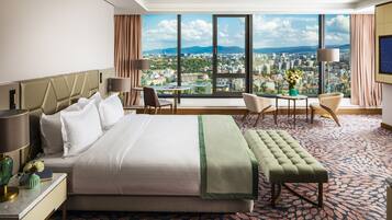 Habitación de hotel con una cama grande, un escritorio, una silla, una mesita y vistas a un paisaje urbano a través de amplios ventanales.